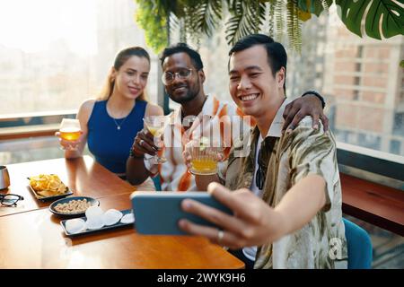 Hugging friends taking selfie when drinking cocktails in rooftop bar Stock Photo