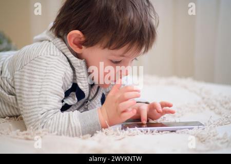 Baby plays with a mobile phone while lying on the bed. The child looks at the phone screen. Kid aged two years Stock Photo