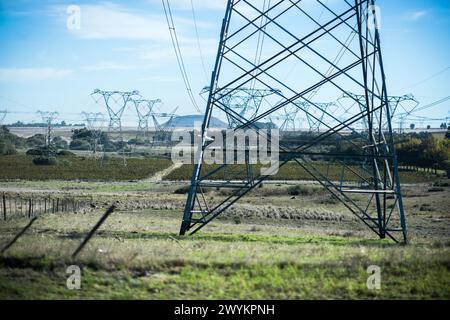 A network of high voltage power lines stretches across a rural landscape, with tall metal towers supporting the electrical cables. The vast open field Stock Photo