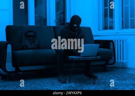 Masked hacker using laptop in dark room Stock Photo