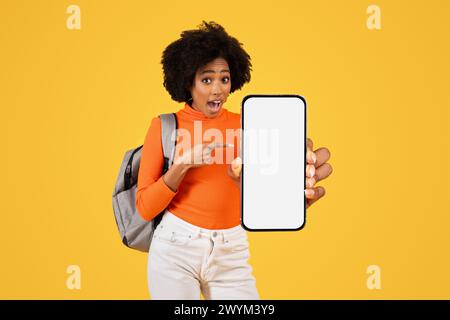 Excited young woman with curly hair, pointing at a blank smartphone screen, dressed in an orange turtleneck Stock Photo