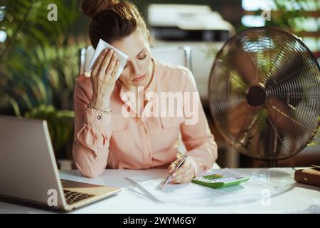 Sustainable workplace. modern middle aged woman employee at work with electric fan, laptop and napkin suffering from summer heat. Stock Photo