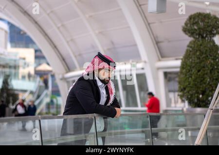 a stylish young man wearing a traditional Arabic headscarf and glasses, leaning against a glass railing in an urban setting. He exudes confidence and Stock Photo