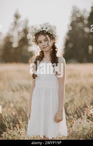 portrait a young girl in a white dress and a floral wreath in the summer field. Boho style. Added a small grain, imitation of film photography Stock Photo