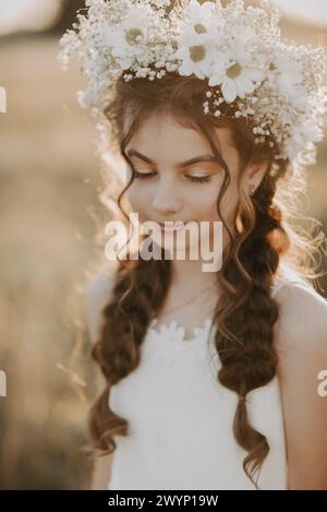 portrait a young girl in a white dress and a floral wreath in the summer field. Boho style. Added a small grain, imitation of film photography Stock Photo