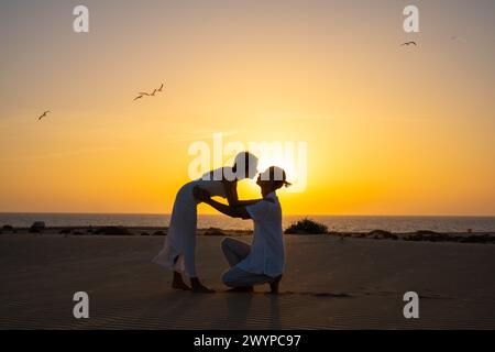 Side view of young couple looking at each other while standing female leaning forward to kneeling male with hair bun against sundown light and orange Stock Photo