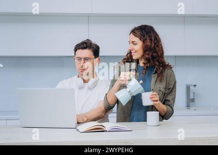 A woman pours coffee for her partner who is focused on work at the laptop, exemplifying a supportive dynamic in a home office setting. A married couple of freelancers work remotely from home, took a coffee break. Stock Photo