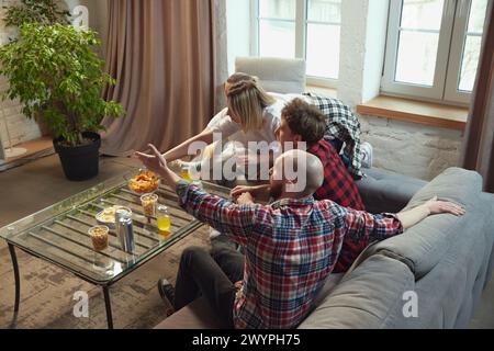 Friends, men and women gathering at home to watch online match translations, eating snacks, emotionally watching game Stock Photo