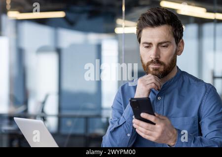 Professional businessman in a modern office setting, intently using a smartphone while working on a laptop. Stock Photo