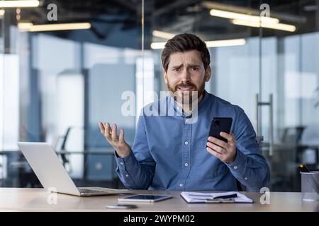 Perplexed young professional with a beard in a blue shirt looking at his smartphone, expressing confusion in a contemporary office setting. Stock Photo