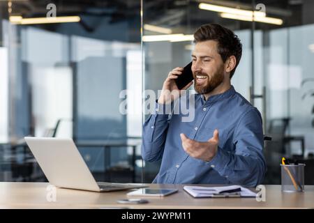A professional man seen mid-conversation on his smartphone, smiling with a laptop and office supplies around him, exemplifying a positive business call. Stock Photo