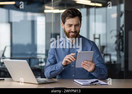 Cheerful bearded man in casual attire enjoying his tablet with a smile at an organized desk in a contemporary office environment. Stock Photo