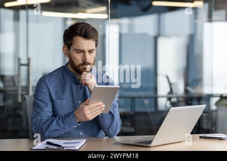 Serious male professional working on a digital tablet with a laptop and notebook by his side in a contemporary office space. Stock Photo