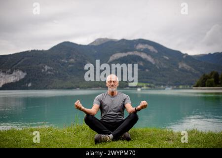 Senior man doing outdoor yoga, meditating by the lake in mountains. Elderly sportsman spending active vacation in nature. Stock Photo