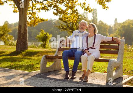 Beautiful stylish elderly couple in love is sitting on bench in city park on warm summer evening. Stock Photo