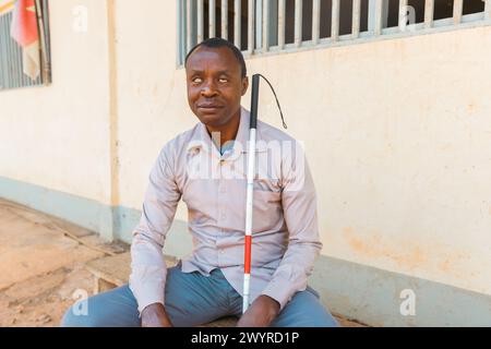 A blind man with a cane sits on a bench. He is wearing a white shirt and gray pants Stock Photo