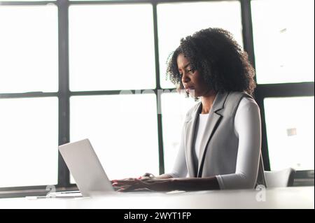 An African-American woman sits and uses laptop, her silhouette framed by the geometric lines of large windows behind her, perfect for themes of modern workspaces and focused professionals. Stock Photo
