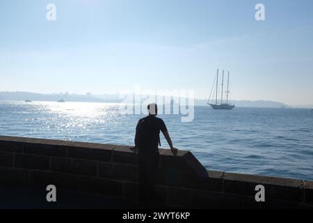 A man looks out across blue water at a three mast schooner on Sydney Harbour on a misty sunny morning from the sandstone wall at Farm Cove Stock Photo
