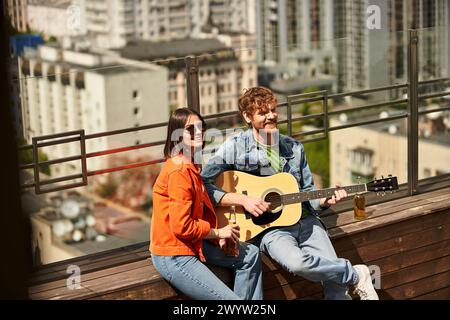 A man and a woman sit on a bench, she holds a guitar while he listens intently. They share melodies under the open sky Stock Photo