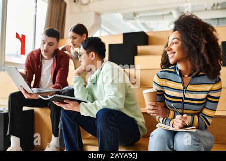 Multicultural students work on laptops while sitting on stairs in an indoor setting, engaging in collaborative learning Stock Photo