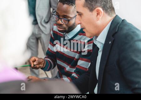 A concentrated pair of professionals, one African man and one Caucasian man, closely examine documents, discussing strategies in a team meeting, their Stock Photo