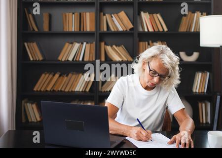 Middle aged senior woman using laptop computer writing notes at workplace. Focused mature old businesswoman work at office making written records doing online work. Mature boss professional worker Stock Photo