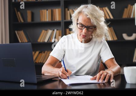 Middle aged senior woman using laptop computer writing notes at workplace. Focused mature old businesswoman work at office making written records doing online work. Mature boss professional worker Stock Photo