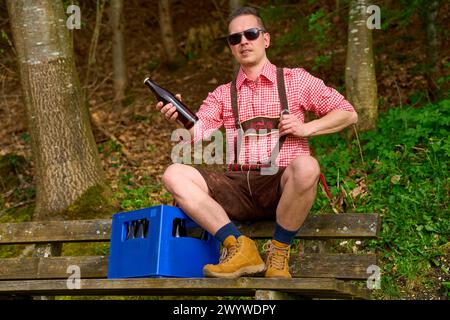 Bavaria, Germany - April 7, 2024: A young Bavarian in lederhosen and traditional shirt on an outing with sunglasses and a crate of beer. Man holding beer bottle *** Ein junger Bayer in Lederhose und Trachtenhemd bei einem Ausflug mit Sonnenbrille und einer Kiste Bier. Mann hält Bierflasche Stock Photo