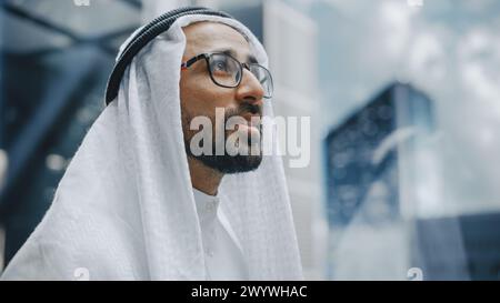 Portrait of a Successful Happy Muslim Businessman in Traditional White Kandura Wearing Glasses Riding Glass Elevator to Office in Modern Business Center. Saudi, Emirati, Arab Businessman Concept. Stock Photo