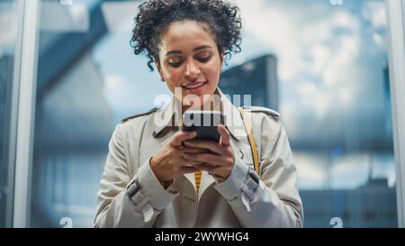 Beautiful Black Female Riding Glass Elevator to Office in Modern Business Center. Successful Manager Smile while Using Smartphone, Write Text Message, Check Social Media and Work Emails in a Lift. Stock Photo