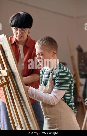 Vertical portrait of smiling boy with disability painting on canvas in art class and wearing apron Stock Photo
