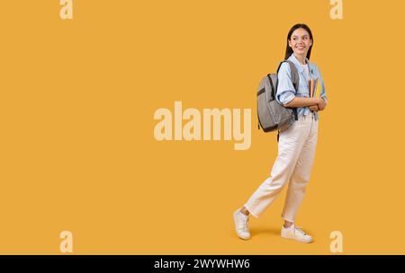 Woman walking with backpack and books on yellow background Stock Photo