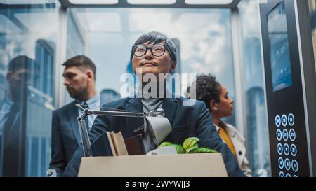 New Employee Coming for a First Day to the Office, Riding in Glass Elevator with Diverse Colleagues in Modern Business Center. Happy Japanese Manager Holding Cardboard Box with Office Accessories. Stock Photo