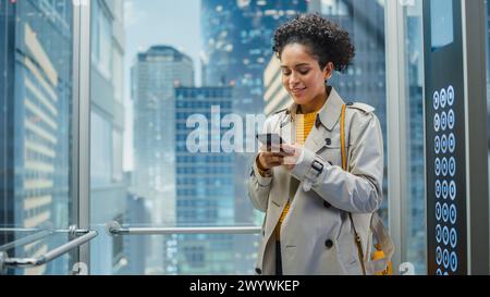 Beautiful Black Female Riding Glass Elevator to Office in Modern Business Center. Successful Manager Smile while Using Smartphone, Write Text Message, Check Social Media and Work Emails in a Lift. Stock Photo