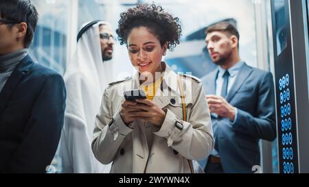 Four Diverse Multiethnic International People Ride a Glass Elevator to Office in a Modern Business Center. Focus on Young Stylish Black Latin Businesswoman Using Smartphone in a Lift. Stock Photo