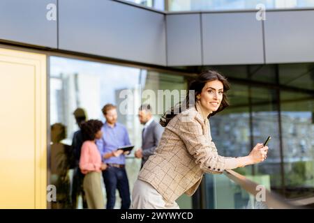 A stylish woman converses on her smartphone, walking past colleagues gathered in casual conversation Stock Photo