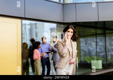 A stylish woman converses on her smartphone, walking past colleagues gathered in casual conversation Stock Photo