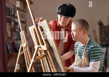 Side view portrait of smiling boy with Down syndrome sitting by easel in art class with teacher assisting Stock Photo