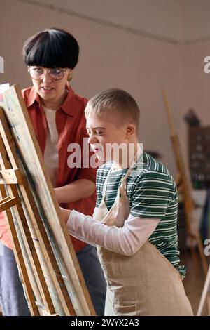 Vertical portrait of smiling boy with disability painting on canvas in art class and wearing apron Stock Photo