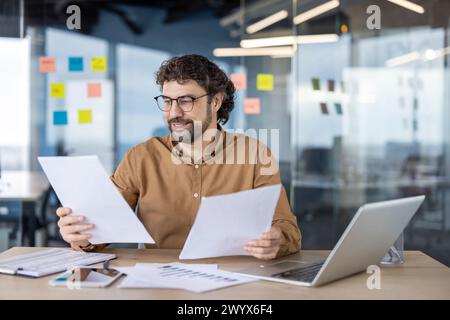 A mature Hispanic man smiling while analyzing papers with a laptop in a modern office setting. Stock Photo