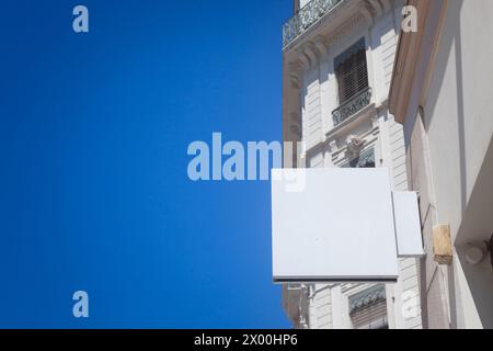 An empty, vibrant white square signboard, prominently displayed and hanging from a metal bracket against a building facade, captures the essence of ur Stock Photo