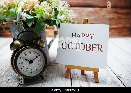 Happy October text message written on paper card with wooden easel and alarm clock with flower in metal vase decoration Stock Photo