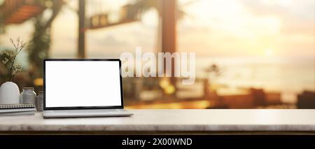 A white-screen laptop computer mockup on a tabletop, with a blurred background of a beachfront hotel lounge or restaurant illuminated by the sunset li Stock Photo