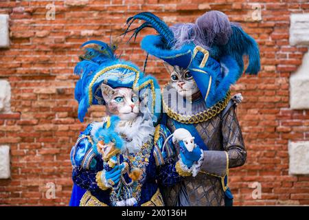 Portraits of two masked persons in creative cat costumes, posing in front of a brick wall, celebrating the Venetian Carnival. Stock Photo