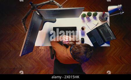 Aerial shot of computer scientist listening music while writing lines of code during remote job shift in home office. IT professional enjoying audio podcast, coding on desktop PC, top down view Stock Photo