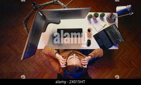 Top down view of technician puts headphones on while fortifying firewalls, teleworking for IT company. Computer scientist listening music and upgrading binary code scripts in home office, aerial shot Stock Photo