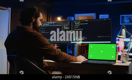 Green screen laptop next to freelancing man doing IT support job from apartment office, coding on PC. Mockup notebook on desk where software engineer writes lines of code on computer at home, camera B Stock Photo