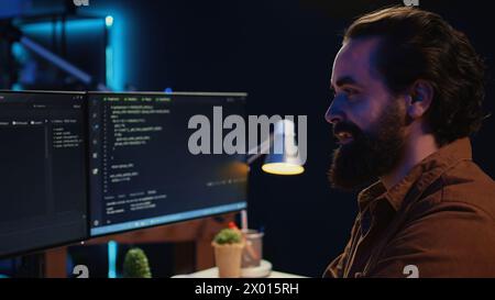 Smiling freelancing developer in home office doing coding on computer at desk. Cheerful IT expert doing contracted job from apartment, writing lines of code at workstation, camera B close up Stock Photo