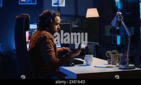 Developer focused on typing complex code in neon lit home office, listening music and developing software application. IT expert wearing headphones, solving coding project tasks on laptop, camera A Stock Photo