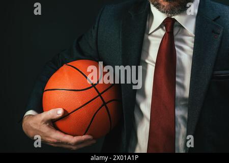 Basketball coach in elegant business suit holding the ball on dark background, selective focus Stock Photo